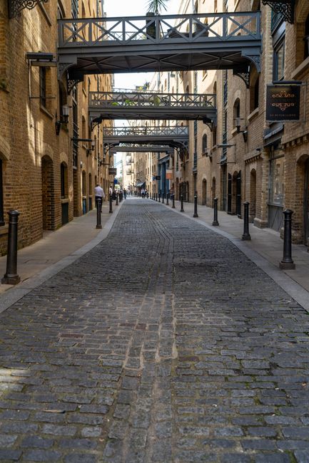 A cobbled street scene in Bermondsey, near Shad Thames, showing two men walking and conversing as they carry a black suitcase between them, indicative of a home relocation or moving process. The stree