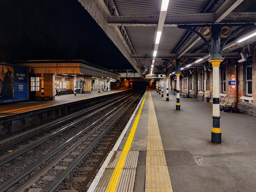 Inside Bermondsey station, the platform features a yellow tactile paving strip along the edge adjacent to multiple railway tracks that extend into the distance. The platform surface is made of concrete and is lined with supporting columns painted in black and beige, supporting the station roof. The sheltered area is illuminated by overhead lights, casting a bright glow on the platform. To the right, there are brick walls with windows and a London Underground roundel sign. In the background, the platform continues with a small waiting area, benches, and a few passengers visible near the station entrance. This scene captures the elements involved in home relocation or furniture transport, with the station environment suitable for loading or unloading items as part of a professional removals service, such as Bermondsey Movers, preparing relocation logistics.