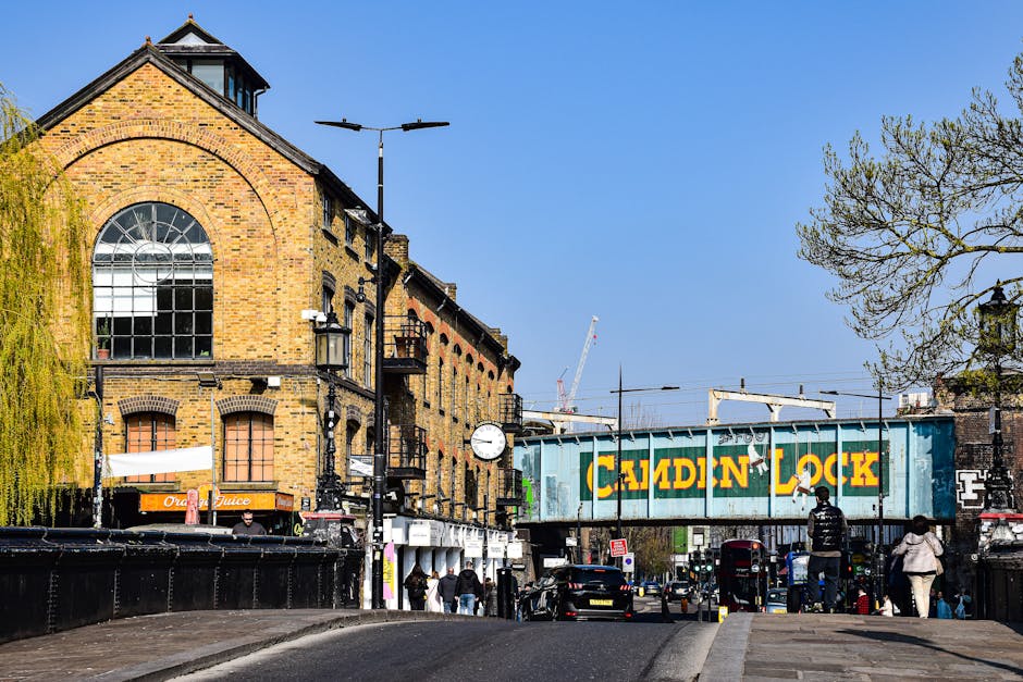 A street scene in Bermondsey featuring a brick building with large arched windows and a clock mounted on a black pole. The building has a sloped roof and is surrounded by trees, including one with drooping yellow-green foliage on the left. The street is bustling with pedestrians walking on the pavement, some carrying bags, and several parked and moving vehicles. An overhead railway bridge with a greenish-blue painted metal structure bearing the yellow text 'CALDYN LOCK' spans across the road. Behind the bridge, construction cranes are visible against a clear blue sky. Streetlights and signage are also present, with the scene capturing the typical urban environment related to home removal and furniture transport processes in the Bermondsey area, close to Bermondsey station.