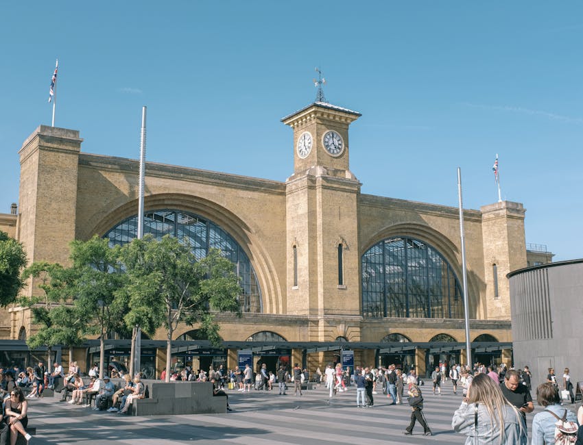 Inside Bermondsey station, the platform features a yellow tactile paving strip along the edge adjacent to multiple railway tracks that extend into the distance. The platform surface is made of concrete and is lined with supporting columns painted in black and beige, supporting the station roof. The sheltered area is illuminated by overhead lights, casting a bright glow on the platform. To the right, there are brick walls with windows and a London Underground roundel sign. In the background, the platform continues with a small waiting area, benches, and a few passengers visible near the station entrance. This scene captures the elements involved in home relocation or furniture transport, with the station environment suitable for loading or unloading items as part of a professional removals service, such as Bermondsey Movers, preparing relocation logistics.