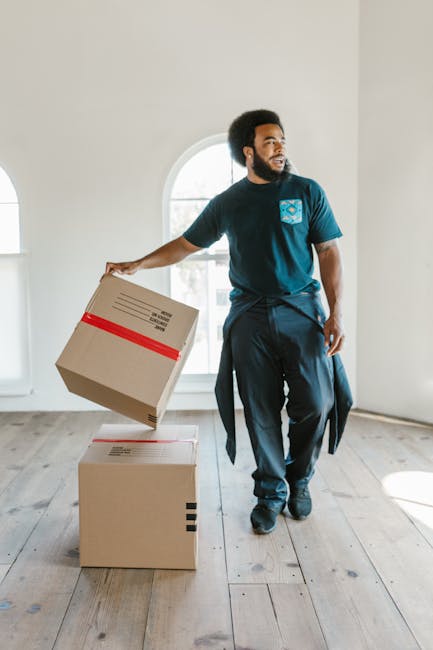 A male mover with dark skin and an afro hairstyle is shown inside an empty room with white walls and a large arched window, which allows natural light to illuminate the space. He is wearing a dark blue uniform with a logo on the chest and is in the process of carrying a cardboard box wrapped with a red packing strap, holding it in his left hand while looking to the side. Two additional cardboard boxes are positioned on the wooden floor nearby – one on top of the other – which are likely filled with household items for home relocation. The room appears prepared for packing and furniture transport, with the smooth wooden flooring visible underfoot. This scene reflects an organized packing and loading process as part of a professional removals service by Bermondsey Movers, focused on house removals and moving logistics.