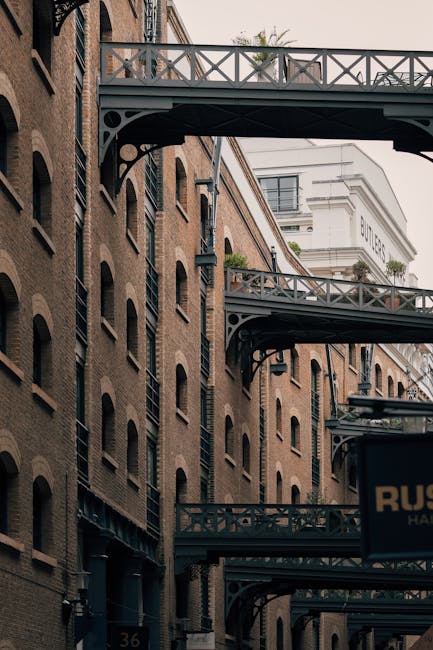 Photograph of a multi-storey brick building with repeated arched windows and decorative metal fire escape walkways connecting different floors, situated along a street in Bermondsey. The image captures the side view of the building during daylight, with a glimpse of modern white structures and a sign reading 'Butlers' in the background. The scene showcases exterior elements often involved in home relocation or furniture transport, including pathways and loading zones, with no visible moving staff or vehicles present. Bermondsey Movers may coordinate moving activities around such buildings for house removals or packing and moving services, emphasizing the urban environment typical of a London residential and commercial area.