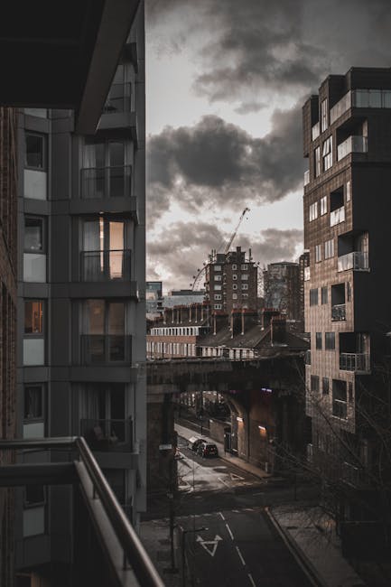 View from a residential balcony showing several modern apartment buildings with large windows and balconies in an urban setting. Outside, a narrow street is visible with parked cars, and an overpass o